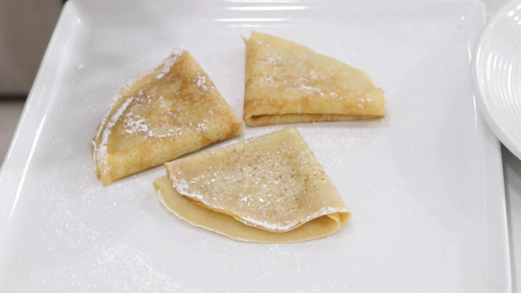 Three gluten free crepes on a white plate ready to be eaten, topped with lemon juice and powdered sugar.