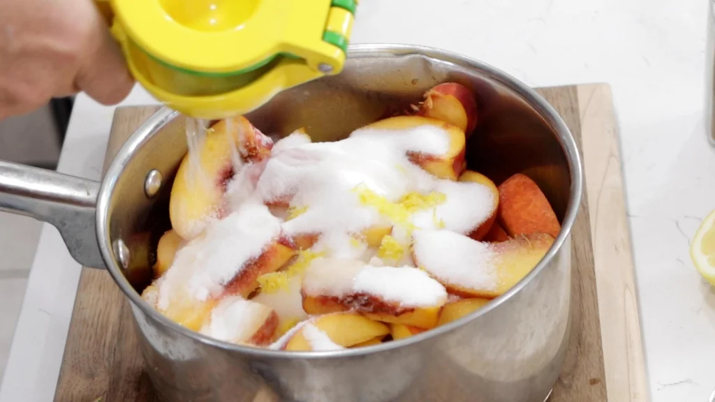 Hand squeezing lemon juice into a metal pot with sliced peaches, sugar, and lemon zest.