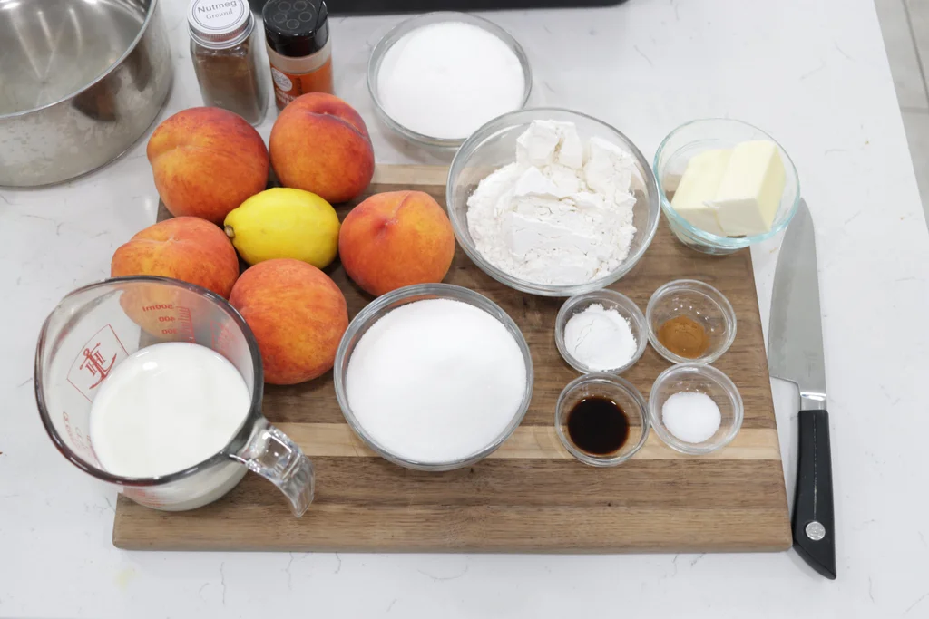 Several ingredients including peaches, sugar, lemon, etc. on a wooden cutting board.