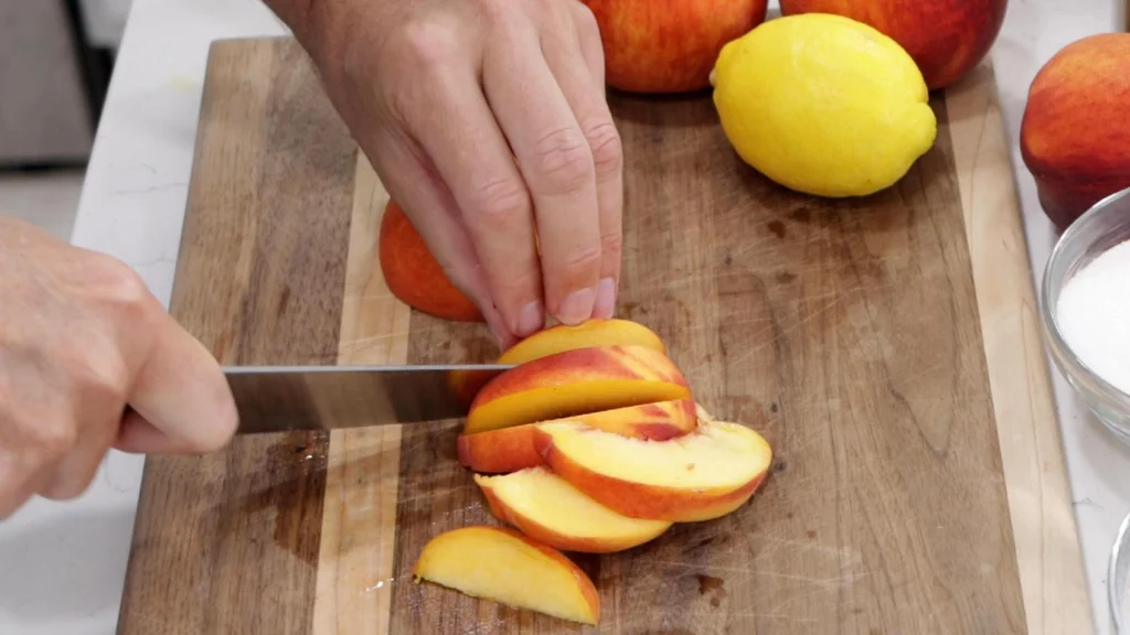 Hand with knife slicing peaches on a wooden cutting board.