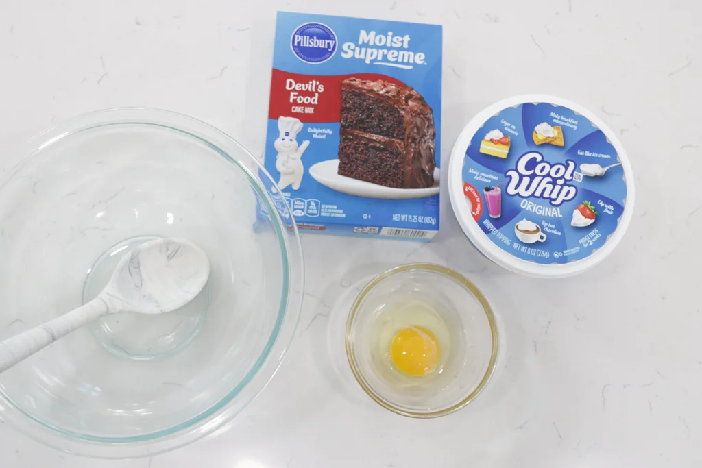 Devil's food cake mix and a tub of Cool Whip on a counter next to a bowl with an egg in it. Devil's food cake mix and a tub of Cool Whip on a counter next to a bowl with an egg in it.