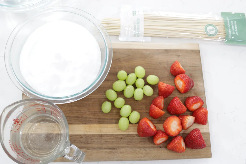 Grapes, strawberries, sugar, and water along with bamboo skewers sitting on a cutting board.