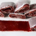 Strawberry fruit leather rolled up in wax paper sitting on a white plate