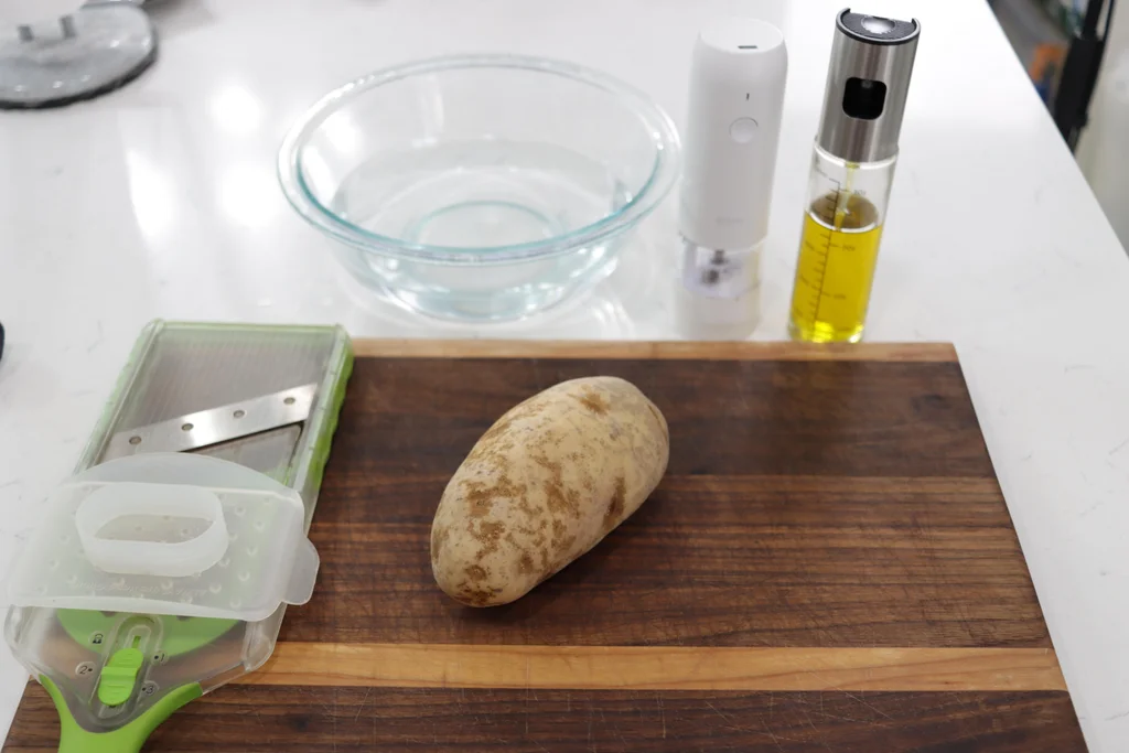 A large Russet potato on a wooden cutting board next to a mandoline and bowl of water. A large Russet potato on a wooden cutting board next to a mandoline and bowl of water.
