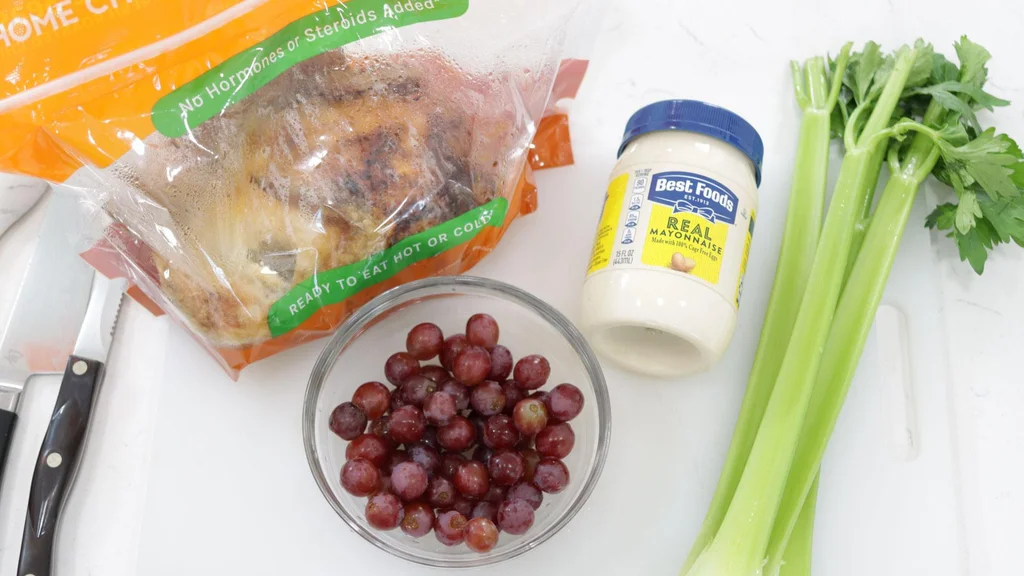 Bagged rotisserie chicken on a white cutting board next to grapes, mayo, and celery. Bagged rotisserie chicken on a white cutting board next to grapes, mayo, and celery.
