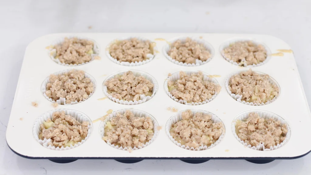 Cream colored muffin pan with paper cups filled with apple muffin batter and crumble topping. Cream colored muffin pan with paper cups filled with apple muffin batter and crumble topping.