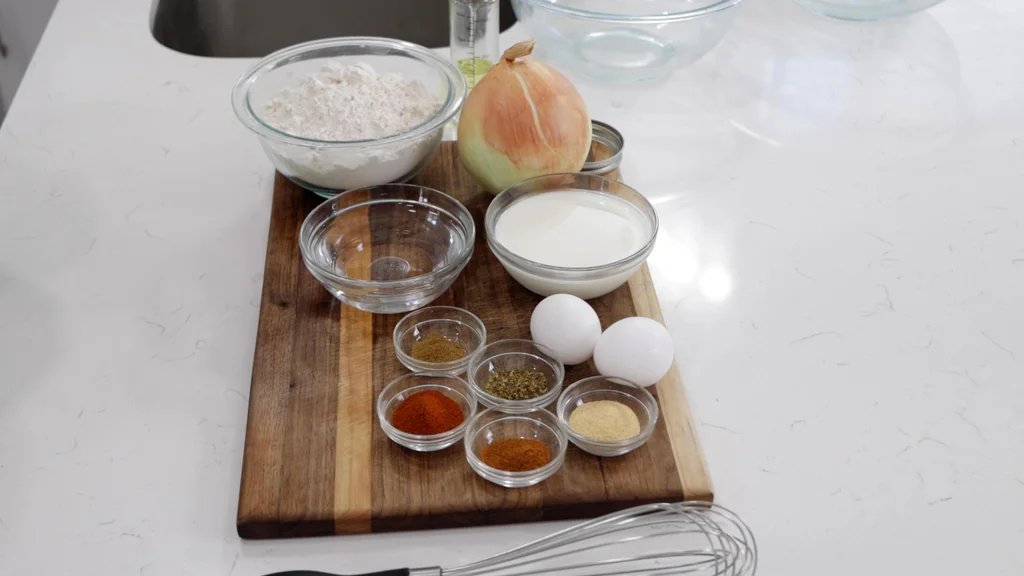 Several ingredients in bowls on top of a wooden cutting board. Yellow onion, milk, flour, eggs, etc. Several ingredients in bowls on top of a wooden cutting board. Yellow onion, milk, flour, eggs, etc.