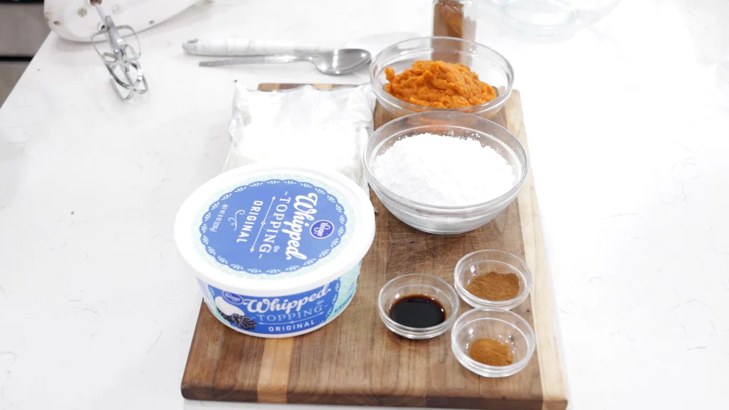 Several ingredients in bowls on a wooden cutting board including cream cheese, whipped topping, canned pumpkin, powdered sugar, etc. Several ingredients in bowls on a wooden cutting board including cream cheese, whipped topping, canned pumpkin, powdered sugar, etc.