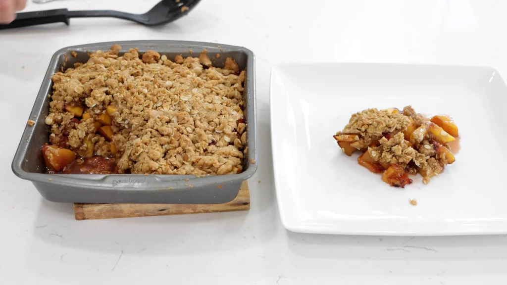 Freshly baked peach crisp still in the pan sitting next to a white plate with a small pile of peach crisp on it. Freshly baked peach crisp still in the pan sitting next to a white plate with a small pile of peach crisp on it.