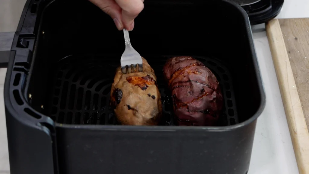 Two sweet potatoes in an air fryer basket, cooked with a fork sticking out of one checking for doneness. Two sweet potatoes in an air fryer basket, cooked with a fork sticking out of one checking for doneness.