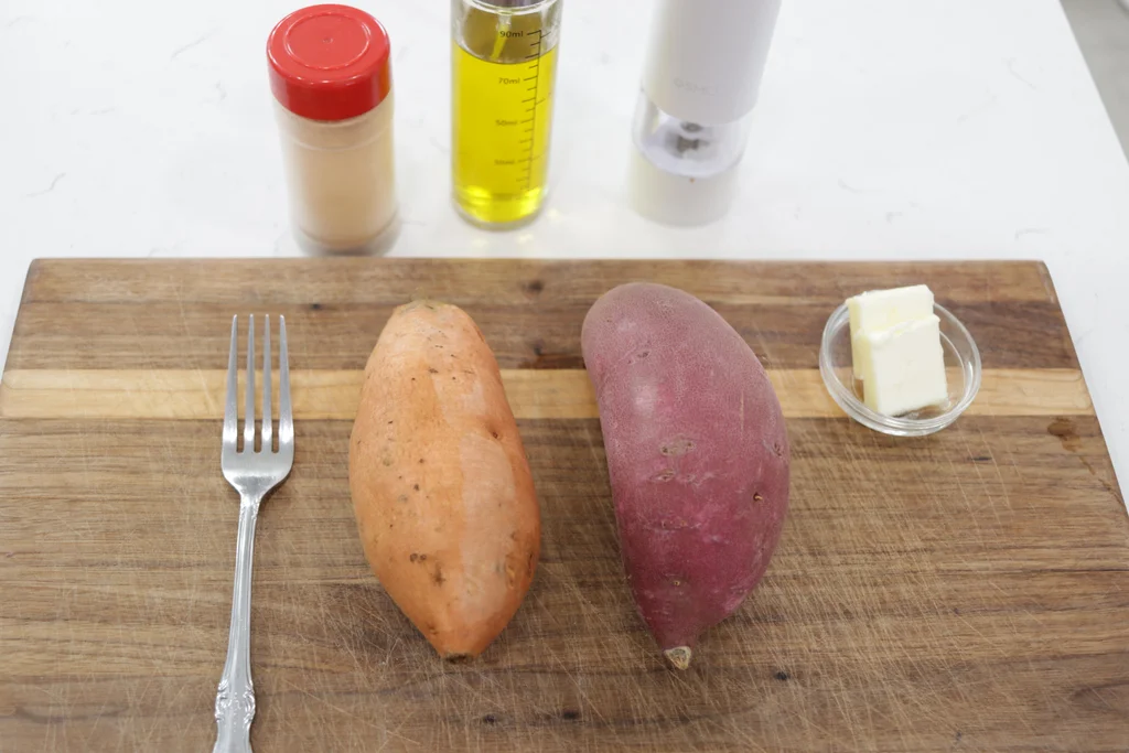 Two sweet potatoes on a wooden cutting board next to butter and a few other things. Two sweet potatoes on a wooden cutting board next to butter and a few other things.