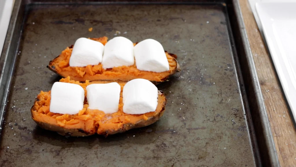 Two sweet potato halves on a sheet pan with large marshmallows on top of each. Two sweet potato halves on a sheet pan with large marshmallows on top of each.