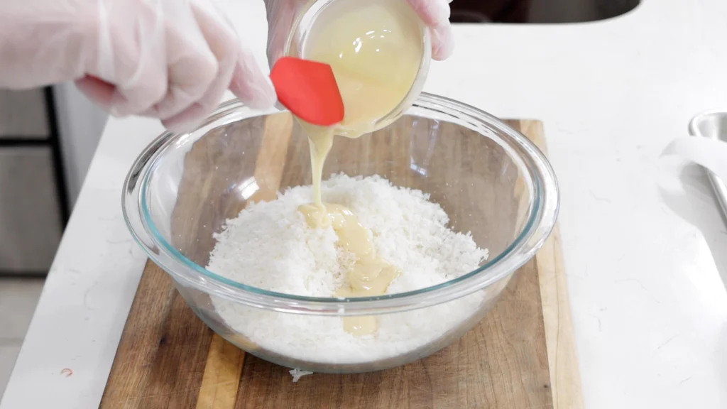 Hands pouring sweetened condensed milk into a bowl of coconut flakes. Hands pouring sweetened condensed milk into a bowl of coconut flakes.