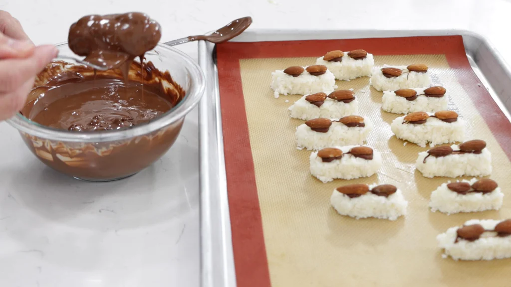 Hand holding a freshly dipped homemade almond joy bar with a fork above a bowl of melted chocolate. Hand holding a freshly dipped homemade almond joy bar with a fork above a bowl of melted chocolate.