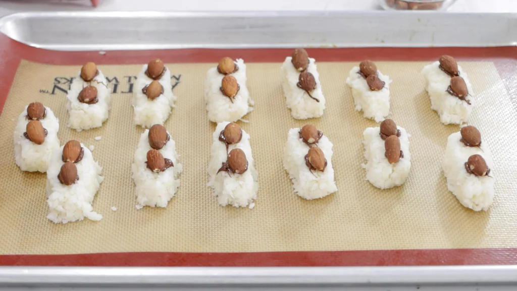 Coconut bars with two almonds on each one, resting on a silicone baking mat inside a sheet pan. Coconut bars with two almonds on each one, resting on a silicone baking mat inside a sheet pan.
