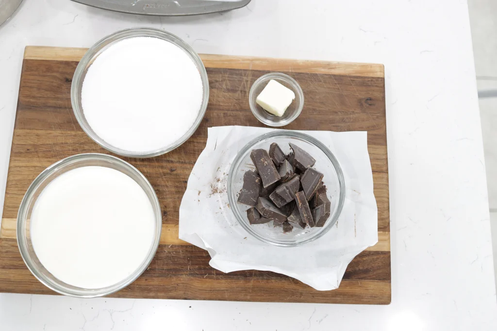 Chocolate, butter, sugar, and cream in bowls on a wooden cutting board. Chocolate, butter, sugar, and cream in bowls on a wooden cutting board.