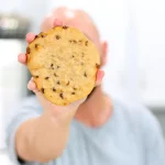Hand holding a protein chocolate chip cookie towards the camera hiding his face.