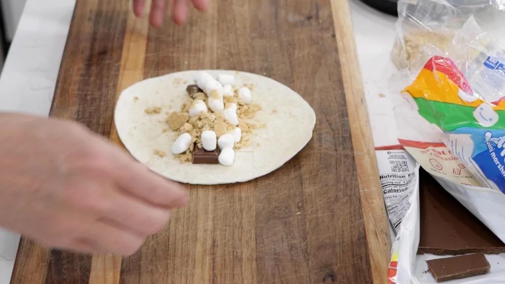 A small tortilla lying on a wooden cutting board with chocolate, mini marshmallows, and graham cracker powder. A small tortilla lying on a wooden cutting board with chocolate, mini marshmallows, and graham cracker powder.
